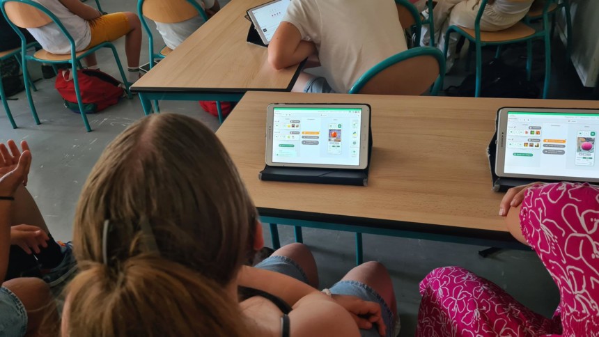 Dans une salle de classe, plusieurs élèves sont assis à des tables. Devant eux, des tablettes affichent un logiciel de programmation visuelle avec des blocs colorés et des images. Deux élèves au premier plan regardent leur écran, tandis que d’autres travaillent également sur leurs tablettes à l’arrière-plan.